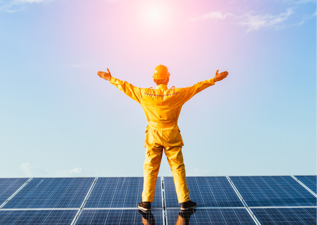 homem vestido de calça e casaco amarelos de costas e capete laranja com os braços abertos ao sol do fundo sobre placas solares